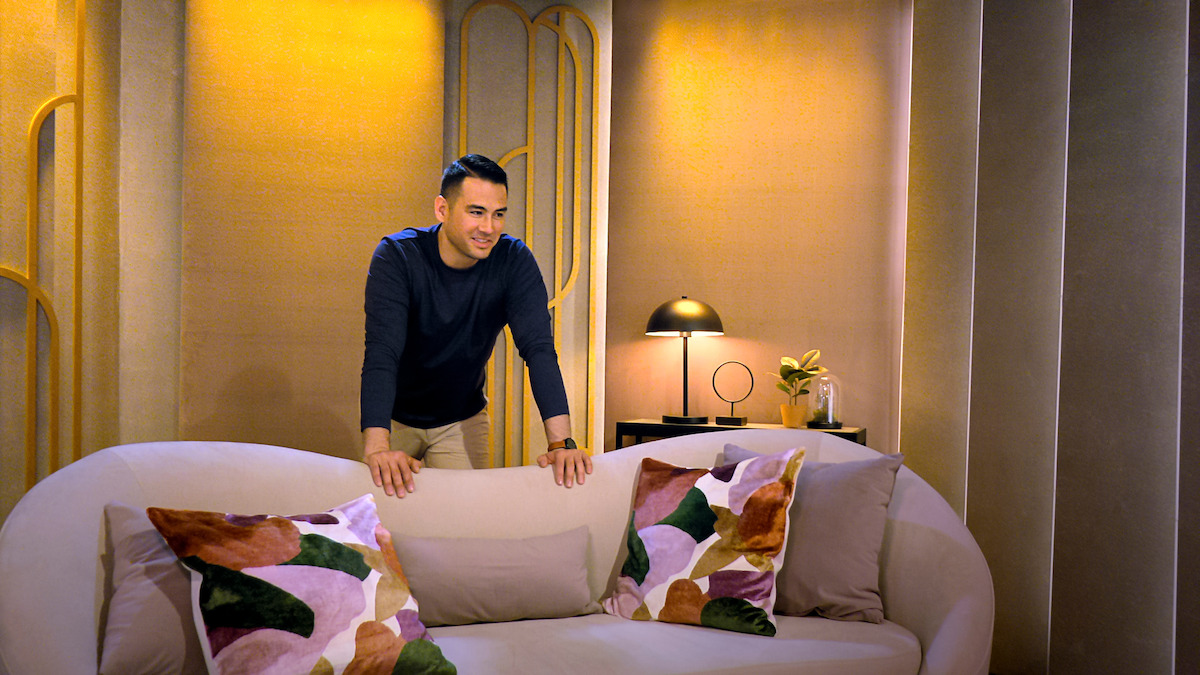 A man stands behind a modern sofa with colorful pillows in a warmly lit, stylish living room featuring gold accents, a side table, and a lamp. The mood is cozy and inviting.