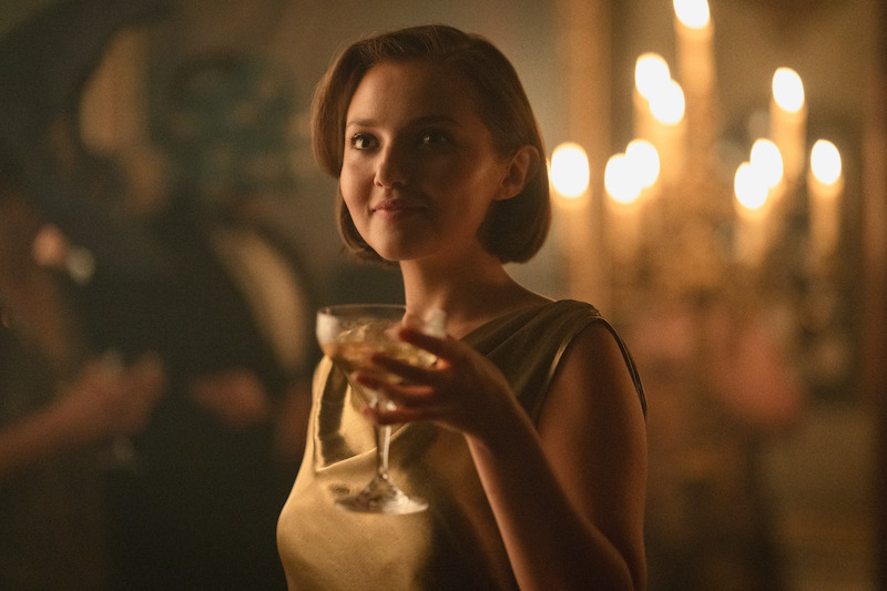 Woman in a gold dress holding a drink at an elegant, dimly lit party with candlelight in the background, creating a warm, sophisticated atmosphere.