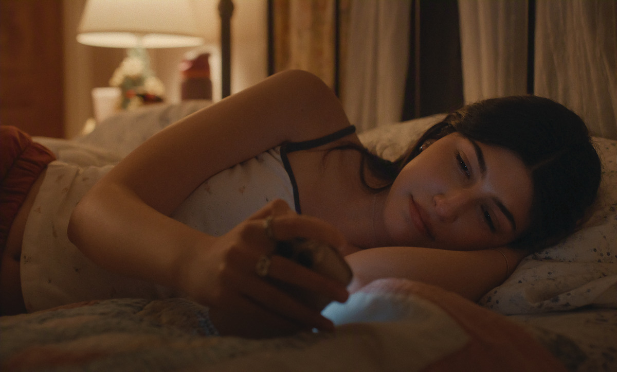Young woman lying on bed in a dimly lit bedroom, holding and looking at a smartphone, with a cozy lamp and decor in the background, suggesting a relaxed evening or nighttime setting.