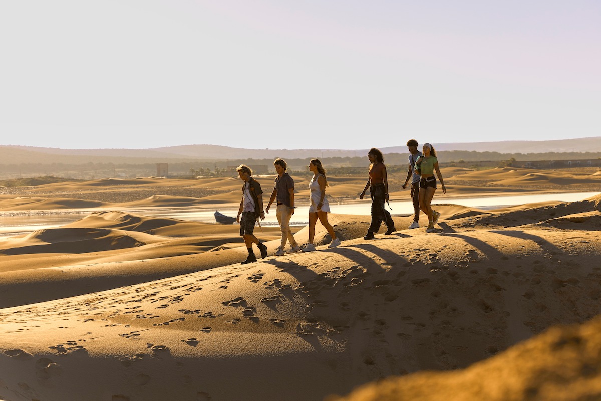 Group of six people walking on sandy desert dunes during daylight, with long shadows and distant water, under a clear sky, surrounded by a wide, open landscape.