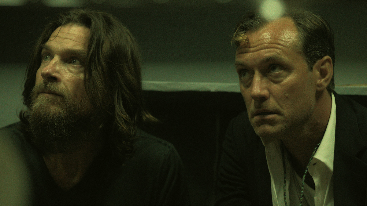 Two men sit closely together in a dimly lit room, one with long hair and a beard wearing casual clothes, the other clean-shaven in a suit, both looking upward with serious expressions, creating an intense, suspenseful atmosphere.