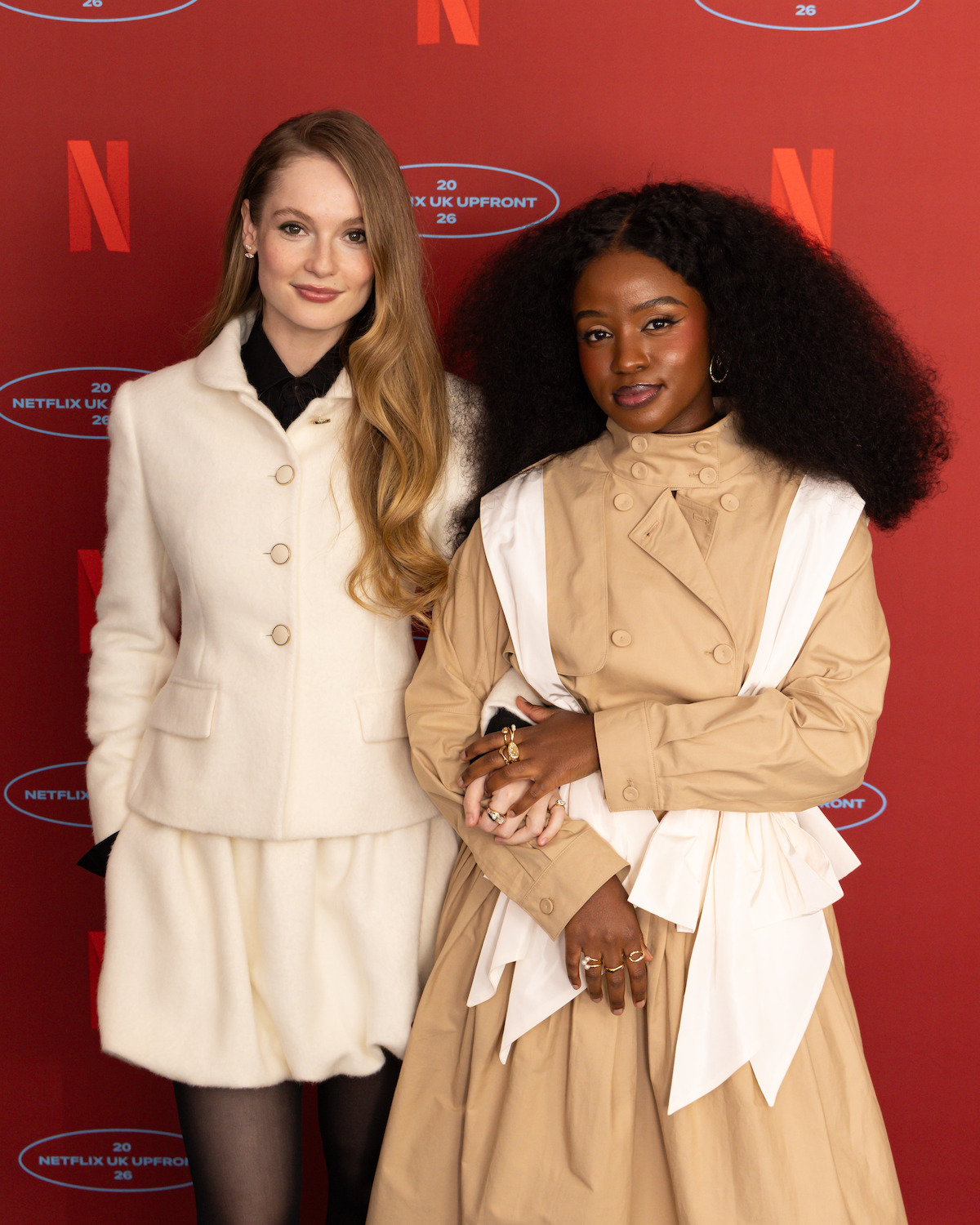 Hannah Dodd and Masali Baduza posing together at a Netflix UK event, standing in front of a red backdrop with Netflix logos. One wears a white outfit, the other wears a beige dress with white accents. Both are smiling in a formal photo setting.