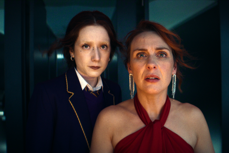 Two people stand indoors with serious expressions, one in a dark school uniform and the other in a red dress with earrings, both illuminated by dramatic lighting, suggesting a tense or emotional moment.