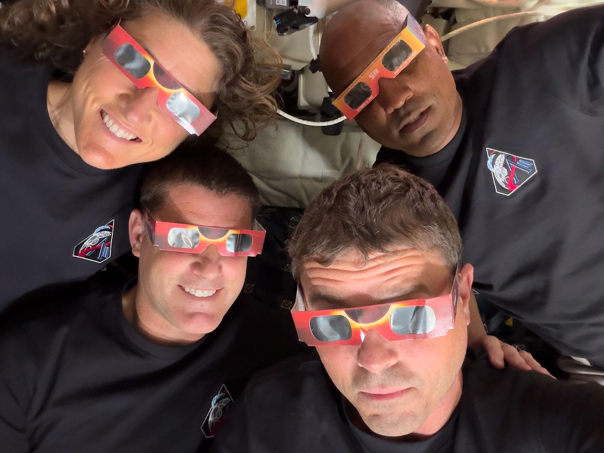 Four people in matching shirts wear solar eclipse glasses and smile, posing closely together indoors in a technical or scientific setting.