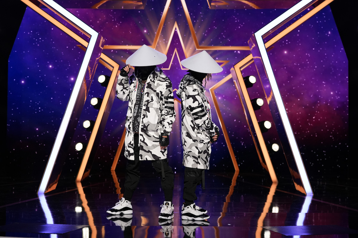 MIV con trajes blancos y negros a juego y sombreros cónicos están espalda con espalda en un escenario con una pantalla de luz violeta en forma de estrella, iluminación geométrica y piso reflectante.