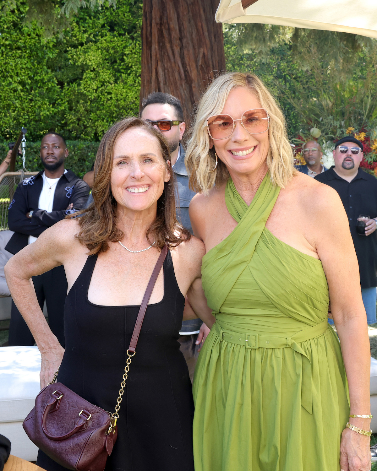 Two women smiling at an outdoor event, surrounded by people and greenery; one wears a black dress, the other a green dress with sunglasses, both appear happy in a sunny garden setting.