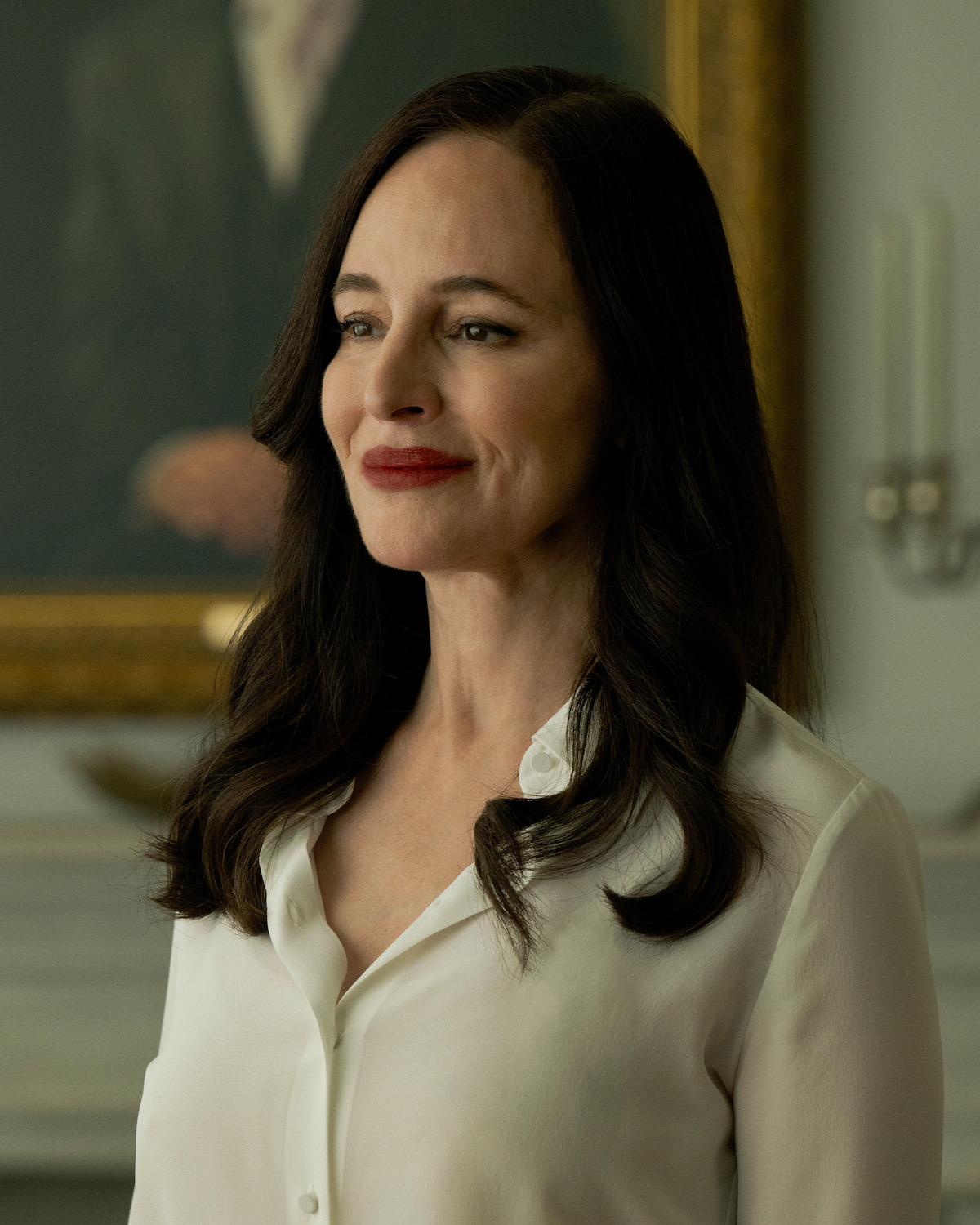 Woman with long dark hair wearing a white blouse stands indoors, smiling slightly. Behind her are candelabras and a framed portrait, indicating an elegant, possibly formal room setting.
