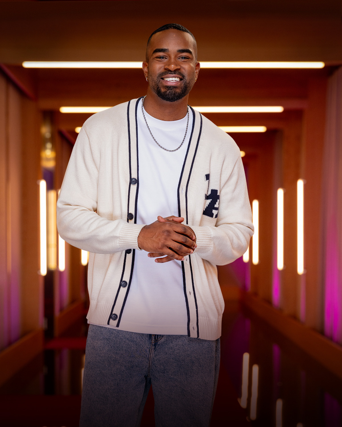 Smiling man wearing a white cardigan and casual jeans stands in a modern, warmly lit hallway with vertical lights and soft ambient colors in the background.