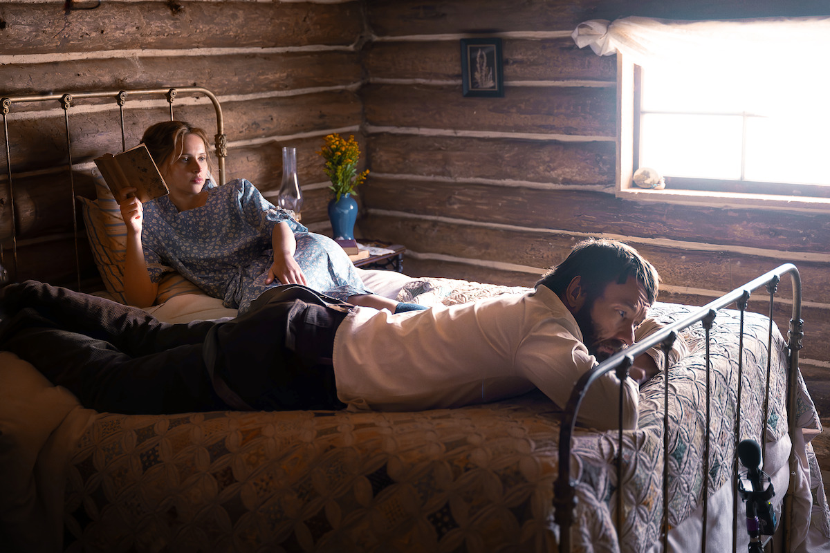A woman reads a book in bed while a man lies beside her in a rustic log cabin room with sunlight through the window, creating a peaceful and intimate atmosphere.