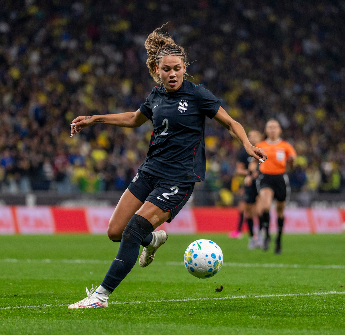 A female soccer player in a black USA uniform controls the ball on a green field, with an official and other players in the background and a large audience in the stadium stands.