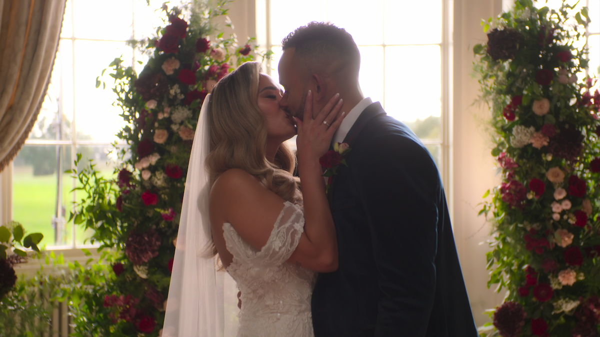 Bride and groom kiss during indoor wedding ceremony, sunlight streaming through large windows, surrounded by floral arrangements, elegant and romantic atmosphere.