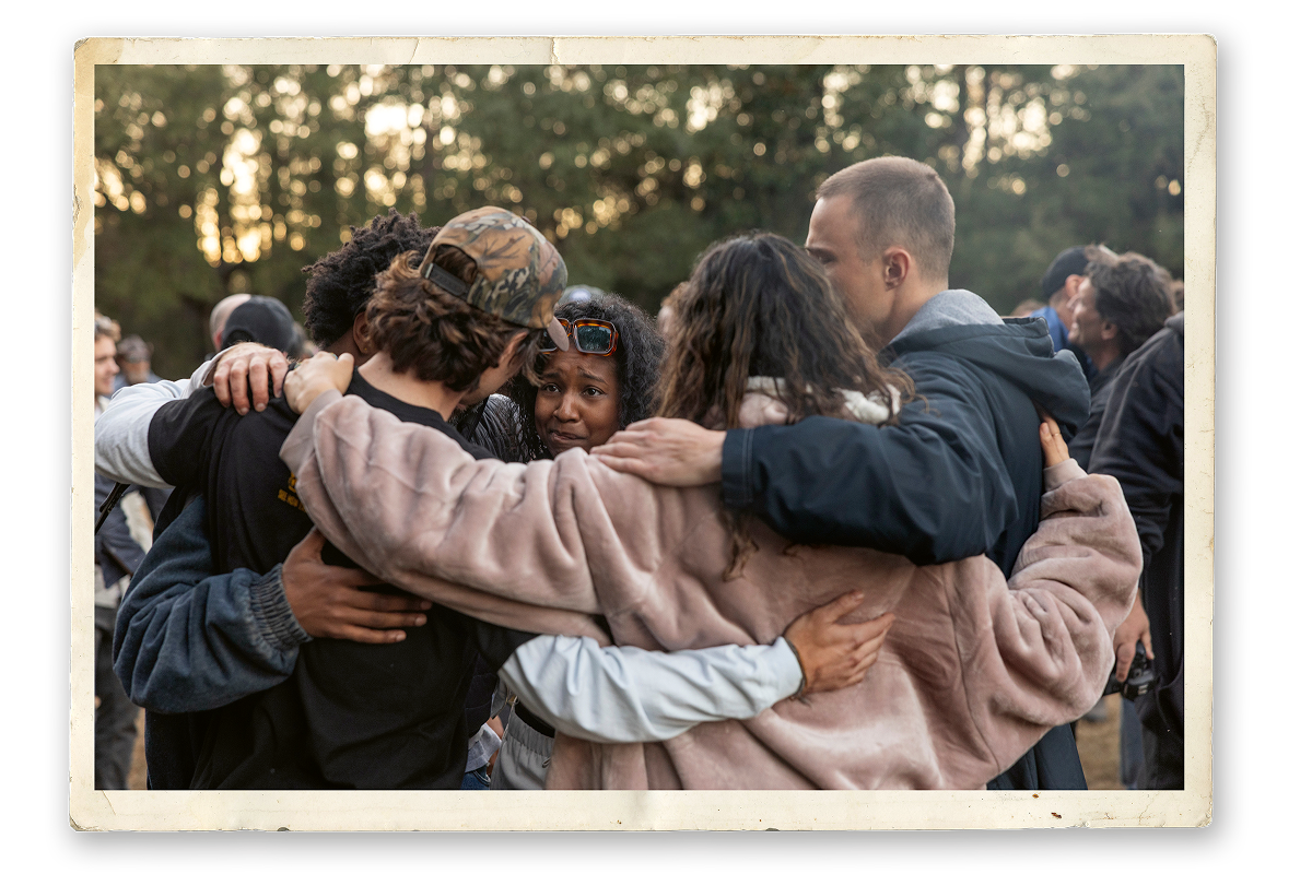 A group of people outdoors, arms around each other in a close huddle, sharing a moment of togetherness at what appears to be a gathering or event, with trees and more people in the background.