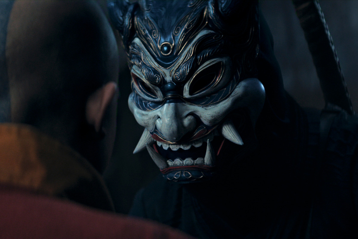 A person wearing a detailed, menacing oni mask confronts a bald figure in a dimly lit, dramatic scene with a dark, mysterious atmosphere.
