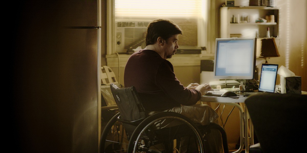 Man in wheelchair working at a desk with computer monitors in a dimly lit home office, window air conditioner and shelves in background.