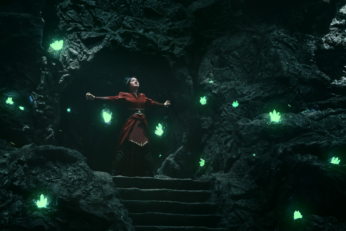 Elizabeth Yu as Azula in a red dress stands with arms outstretched in a dark cave entrance, surrounded by glowing green crystals on rocky walls, with stone steps leading up to her.