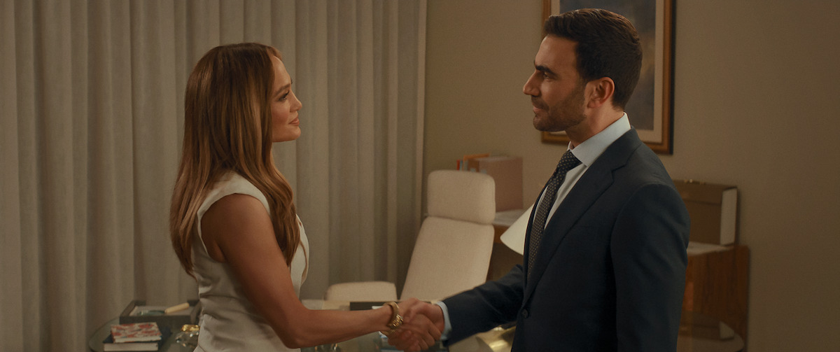 A woman in a white dress and a man in a suit shake hands in a modern office with a desk, chair, and curtain in the background, suggesting a professional meeting or agreement.