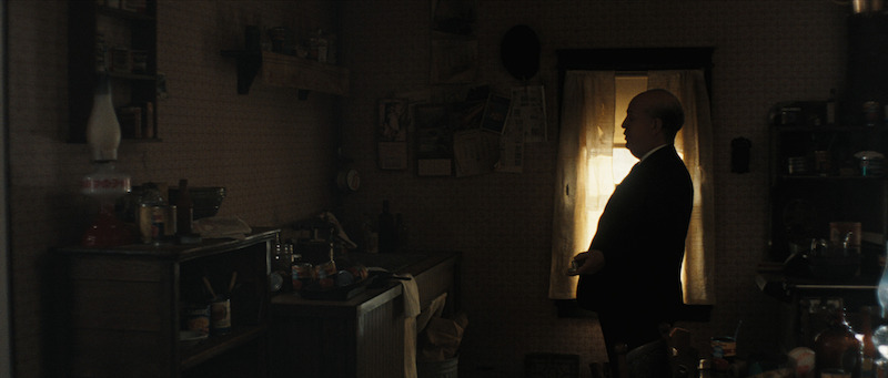 A man in a suit stands in a dimly lit, cluttered kitchen, facing a window with light streaming through the curtains.