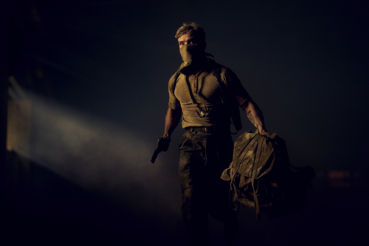 A masked man with a gun and a duffel bag walks through a dark, smoky environment with a beam of light shining in the background, suggesting a tense or action-packed setting.