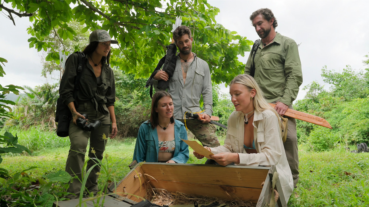 Five people outdoors in a green, forested area gathered around an open wooden chest; one person is holding papers and discussing findings as others watch attentively, with relaxed, adventurous expressions.