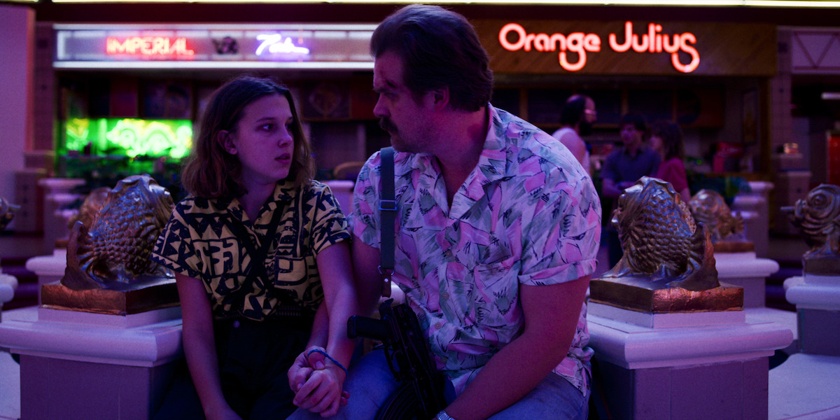 A man and a teenage girl sit closely on a mall bench, surrounded by neon lights and storefronts.