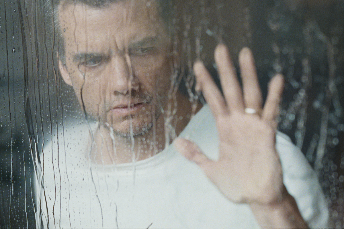 Man in white shirt standing indoors, looking out somberly through a rain-covered window, touching the glass with his hand, with drops of water and reflections creating a moody, introspective atmosphere.