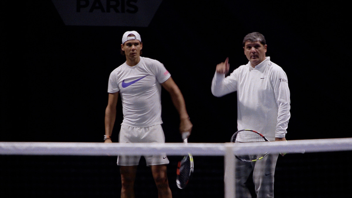 Two men standing on a tennis court, both holding rackets, one in sportswear and the other in casual clothes, with a net in the foreground and a dark background. Indoor setting, possibly during a training session or event.