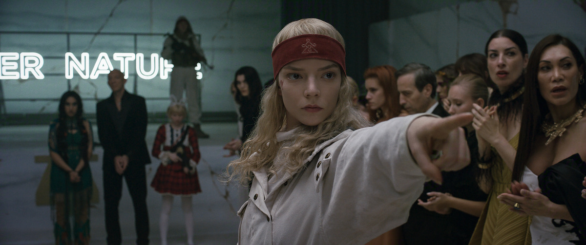 A blonde woman wearing a red headband points with intensity in a modern room with marble walls, neon lights, and a group of stylishly dressed onlookers watching her action closely.