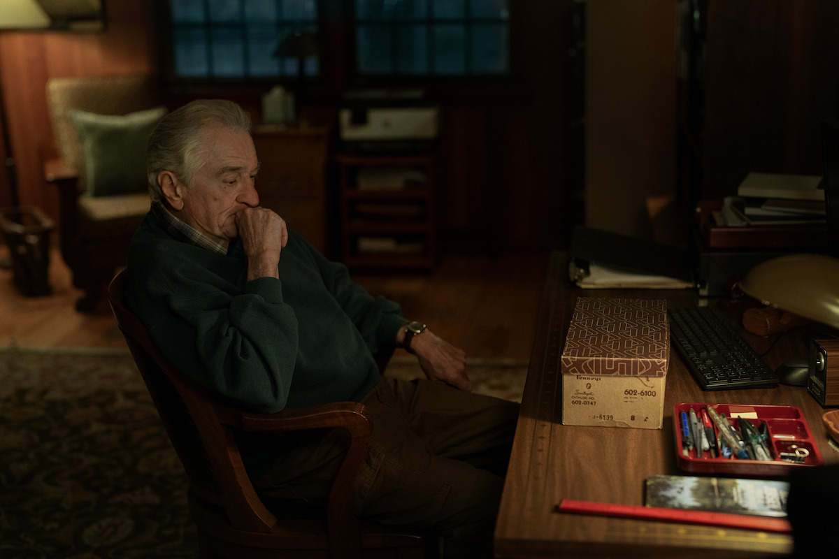An elderly man sits thoughtfully at a desk in a dimly lit home office, surrounded by stationery, a computer keyboard, a lamp, and a box, with shelves and a large window in the background.