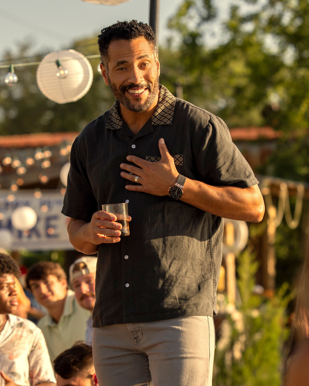 Marland Burke as Mike Carrera, holding a drink and making a speech outdoors at a daytime gathering, with festive lights, paper lanterns, and people seated in the background, surrounded by greenery.