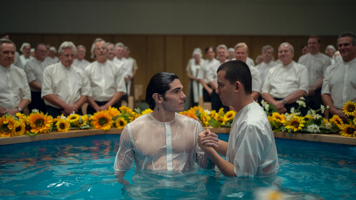 Two men in white shirts stand in a baptismal pool surrounded by sunflowers, with a group of people in white shirts and blouses watching in the background, in a brightly lit indoor setting.