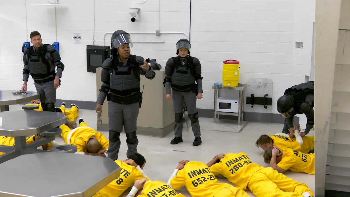 Correctional officers in tactical gear supervise and restrain inmates in yellow uniforms lying face down on the floor of a brightly lit prison common area with tables and a water dispenser visible.