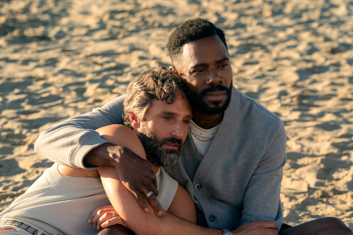 Two men sit close together on a sandy beach, embracing each other for comfort and support under warm sunlight, creating an intimate and serene atmosphere.
