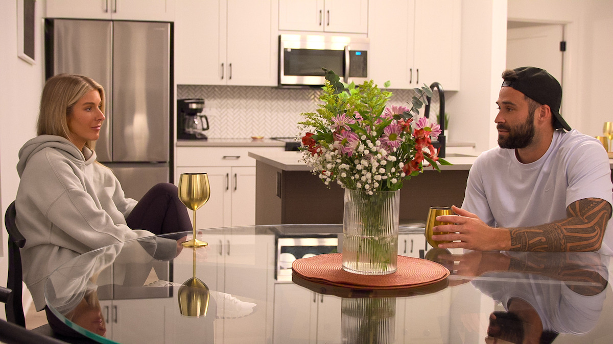 Ashley Carpenter and Alex Henderson sit at a glass dining table with gold wine glasses and a bouquet of flowers in a vase, having a conversation in a modern, bright kitchen with white cabinets and stainless steel appliances.