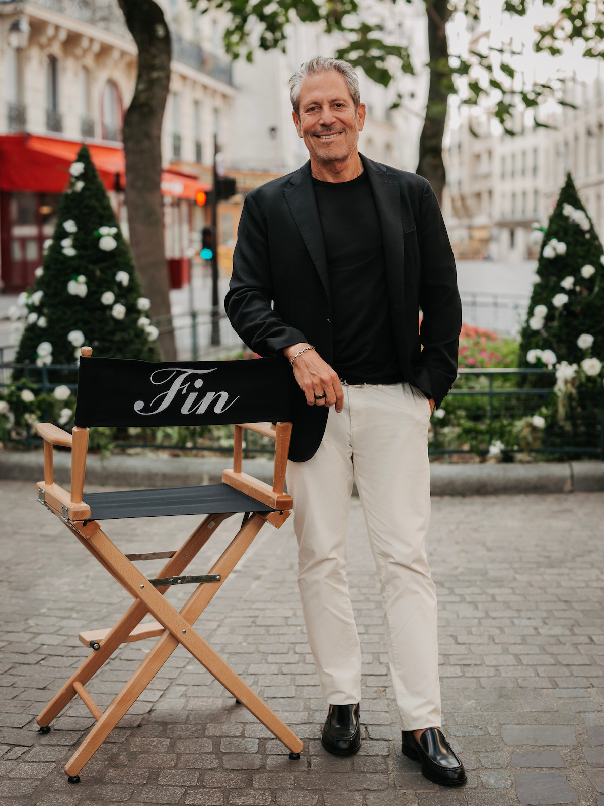 Smiling man in black blazer and white pants stands next to a director's chair labeled "Fin" on a cobblestone street, with greenery, flowers, and city buildings in the background.