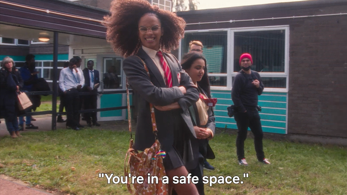 Two students smiling and walking together outside a school building with other people in the background; the environment is casual and friendly. The subtitle reads "You're in a safe space."