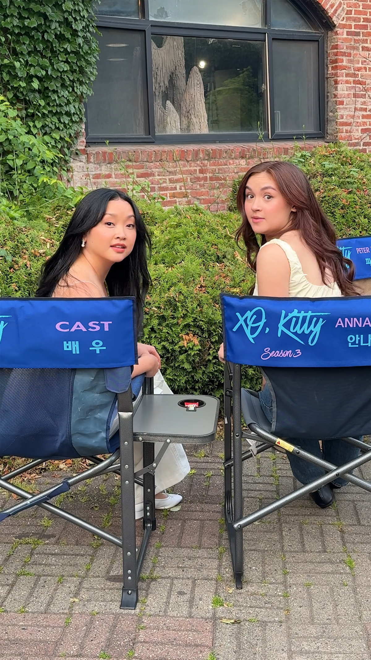 Two women sitting outdoors on blue director’s chairs labeled for the cast of XO, Kitty Season 3, with greenery, brick wall, and a large window in the background, both looking back over their shoulders at the camera.