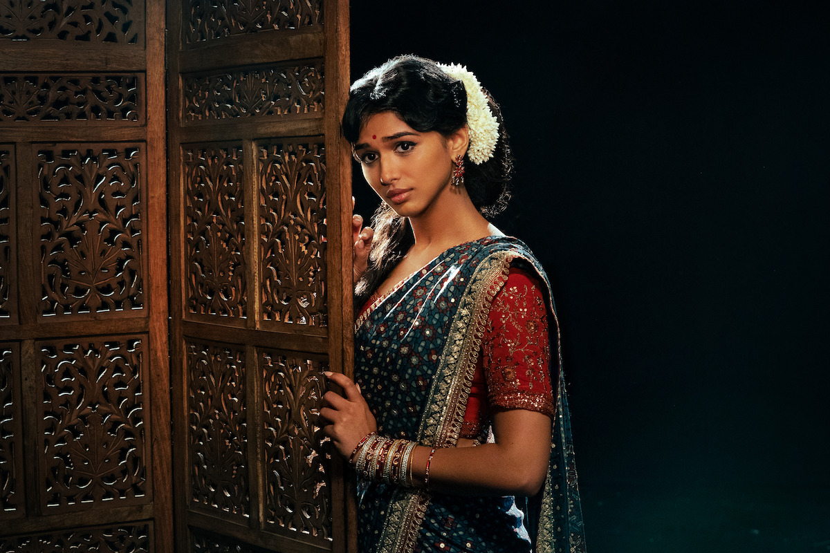 Woman in traditional Indian attire with flowers in her hair stands beside a wooden carved screen in a dimly lit room, looking thoughtfully at the camera.