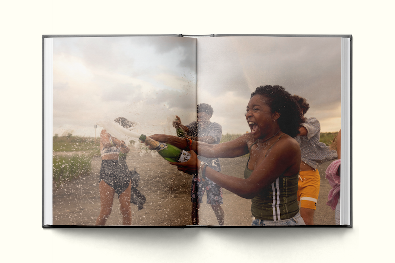 A joyful group of young people outdoors, one woman in front sprays champagne, laughing, with friends in the background, grassy field, cloudy sky, captured in an open photo book.