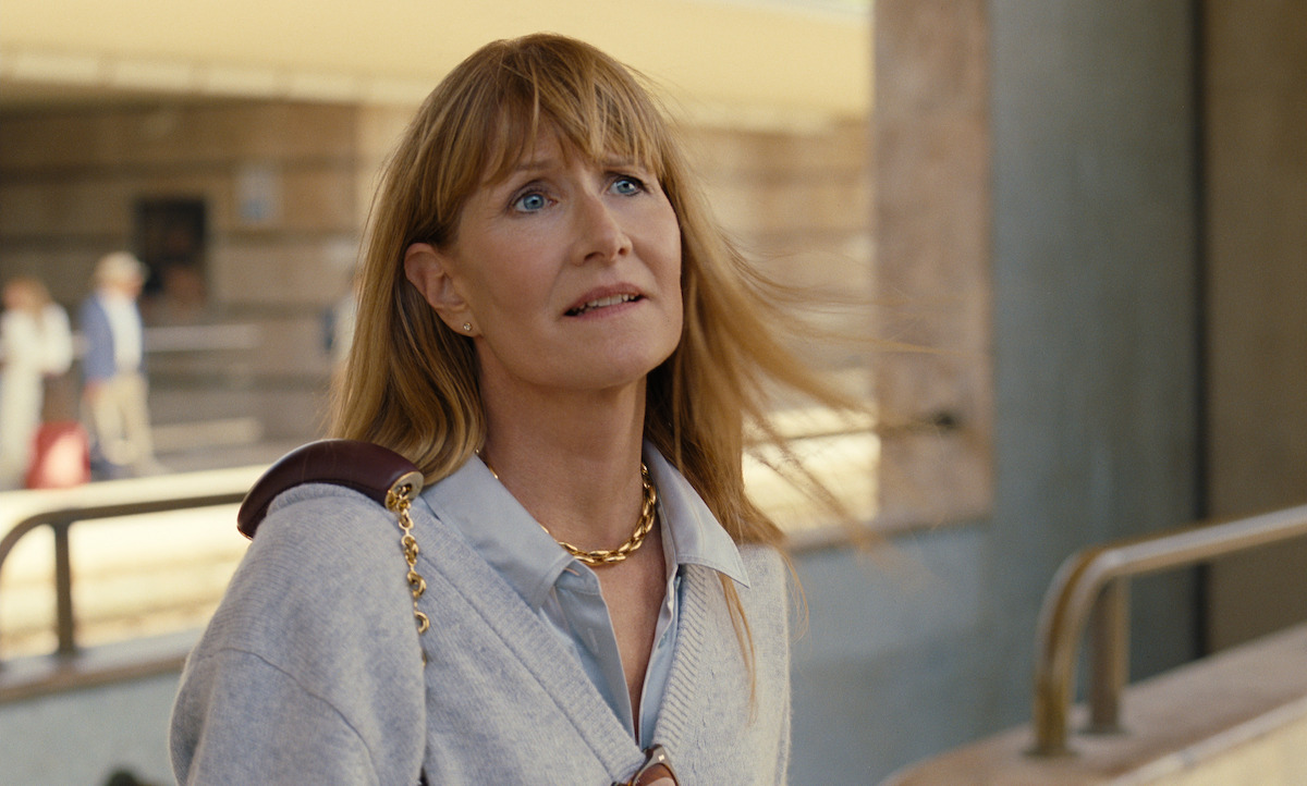 A woman with blonde hair and a gold chain necklace stands outdoors near a building, looking thoughtful and slightly concerned, with wind blowing her hair. The setting appears urban and the mood is contemplative.