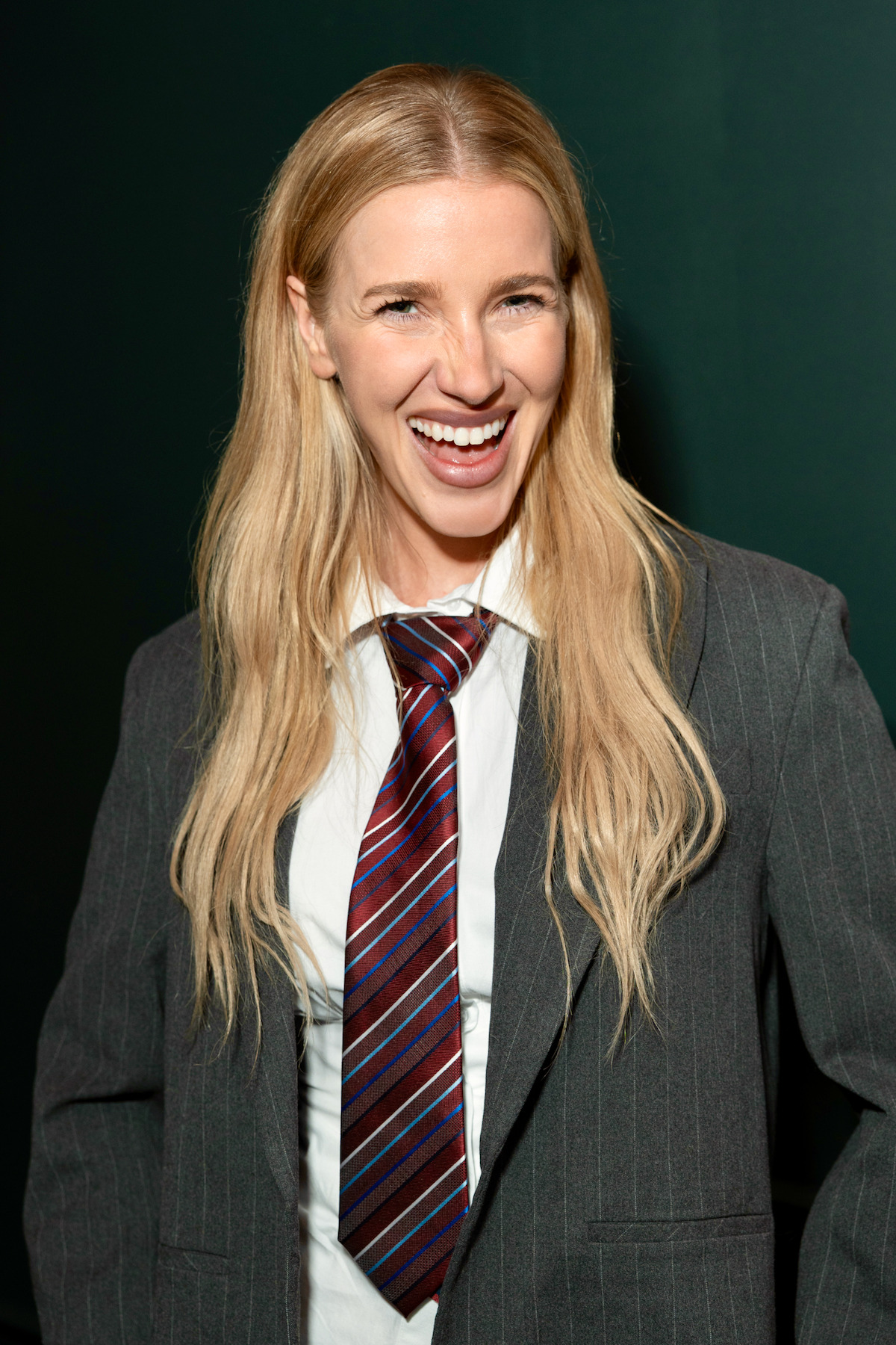 Smiling woman with long blonde hair wearing a grey pinstripe suit and striped tie, standing indoors against a dark green background.