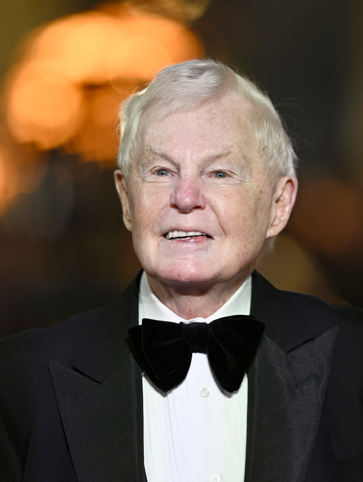 Elderly man in formal attire with black tuxedo and bow tie, smiling at an indoor event with blurred warm lights in the background.