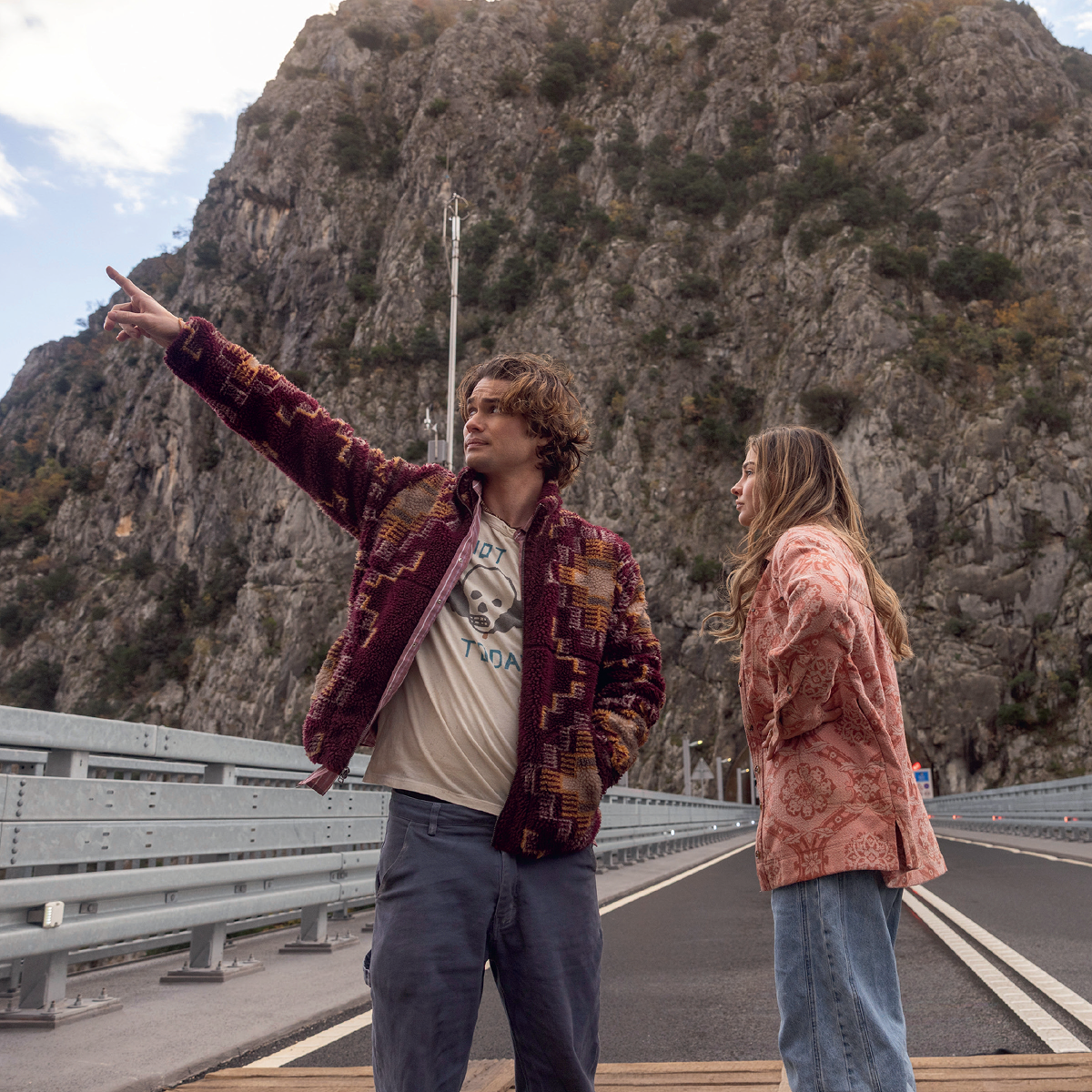 Two people standing on a road bridge by a mountain, one pointing into the distance while talking to the other, both dressed casually, surrounded by rocky landscape and autumn foliage.