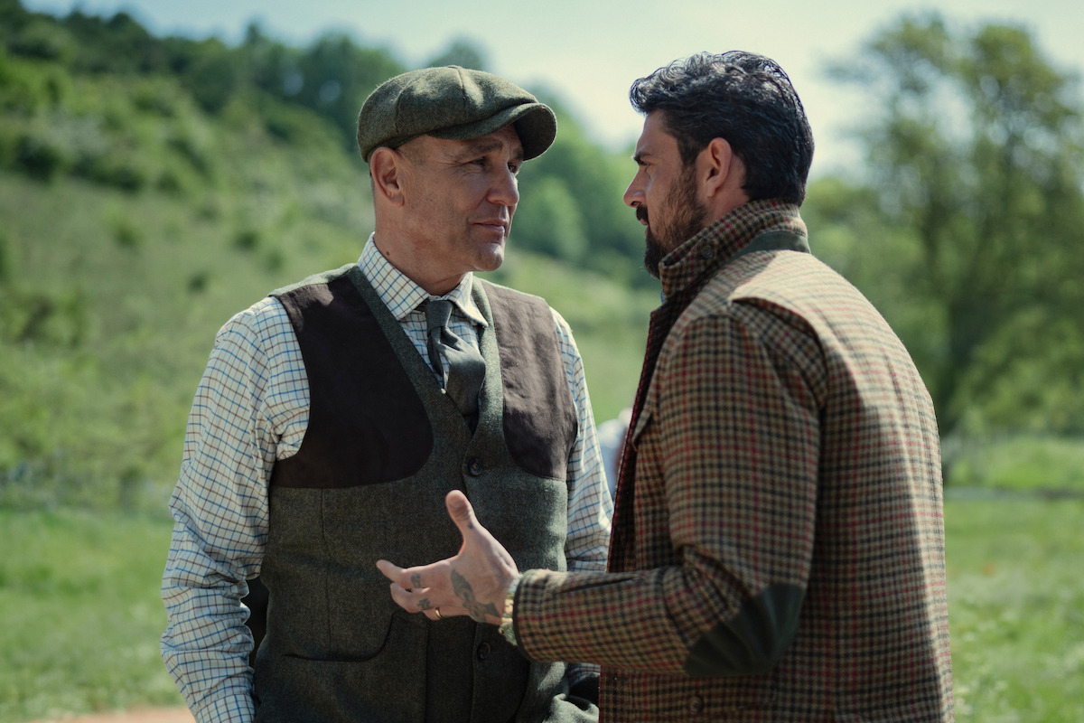 Two men stand outside in a rural setting, dressed in country-style jackets and hats, engaged in a serious conversation with greenery and trees in the background.