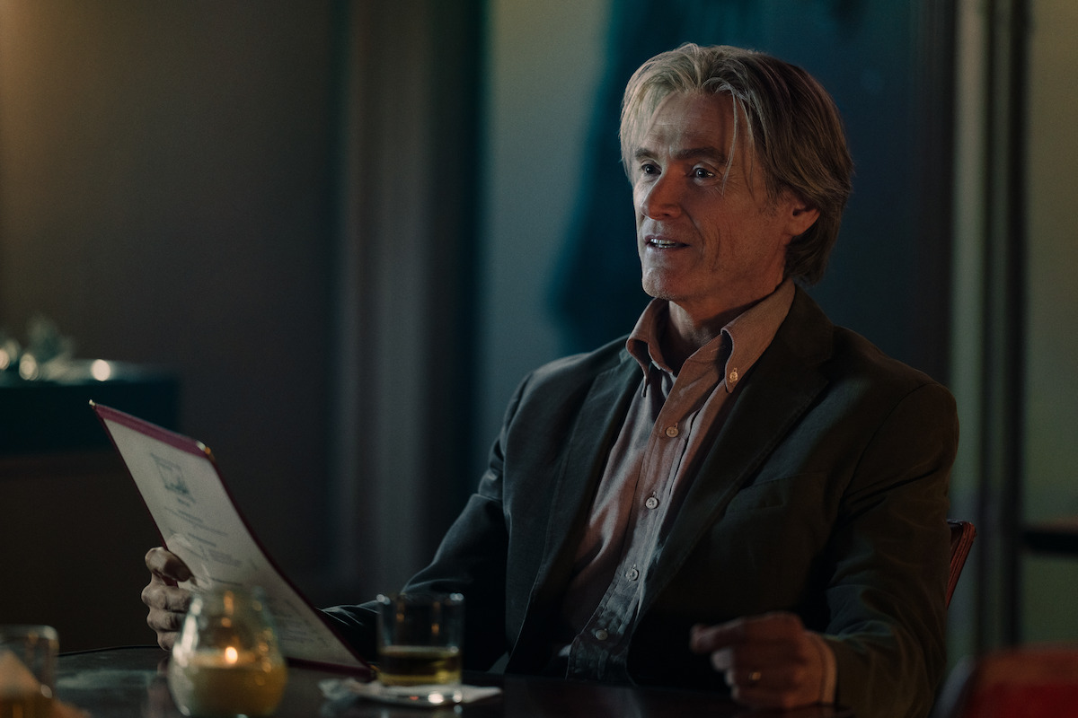 Older man in a dimly lit restaurant, holding a menu and sitting at a table with a drink and candle, dressed in a suit, appearing engaged and content in a relaxed, intimate setting.