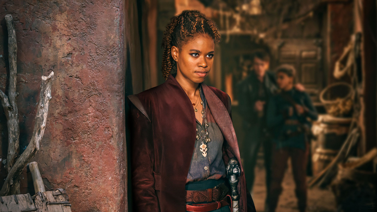 A determined woman in fantasy attire stands by a weathered wall in a rustic, dimly-lit environment with two figures blurred in the background, creating a tense and adventurous mood.