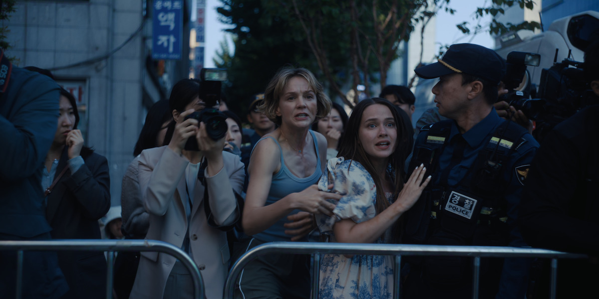 A distressed woman and a girl are held back by a police officer amid a crowd and media with cameras on a city street, separated by a barricade, during what appears to be a tense or chaotic public event.