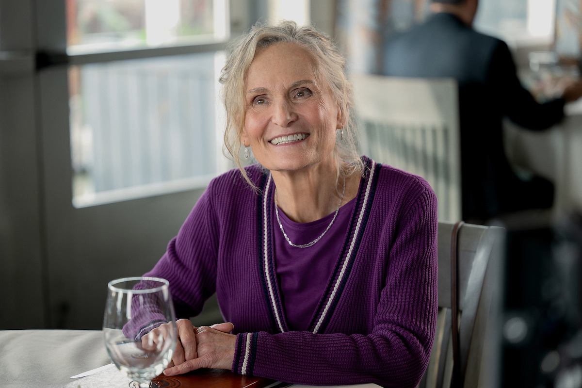 Beth Grant as Barb, with light hair in a purple sweater sits at a table in a bright, modern restaurant, holding a glass, with another person blurred in the background.