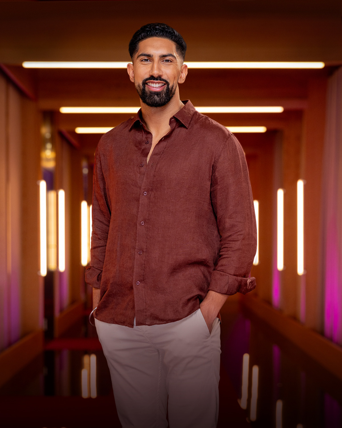 Man with short dark hair and beard wearing a brown shirt and light pants, standing in a modern hallway with warm lighting and glowing vertical light fixtures in the background.