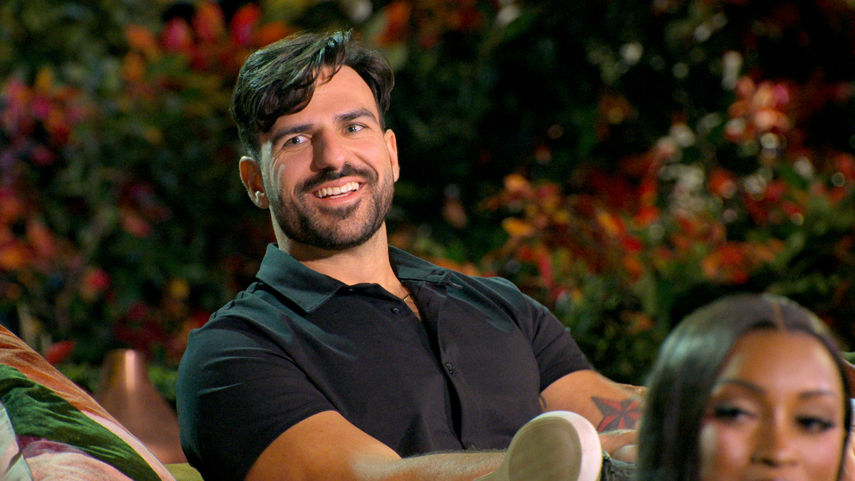 A man in a black shirt sits on a couch outdoors, smiling and relaxed, with vibrant greenery and colorful plants in the background, creating a cheerful, casual atmosphere. Another person is partially visible in the foreground.