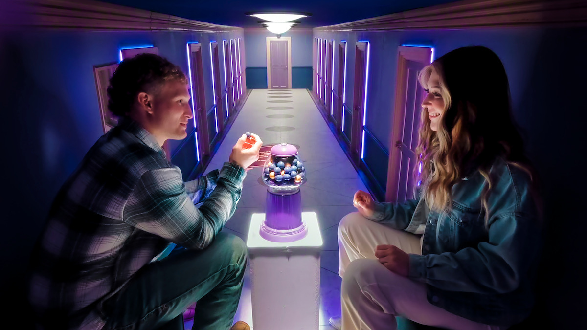 Two people sit facing each other in a hallway with glowing purple light, next to a gumball machine. The hallway has multiple doors and a bright light at the end, creating a surreal atmosphere.